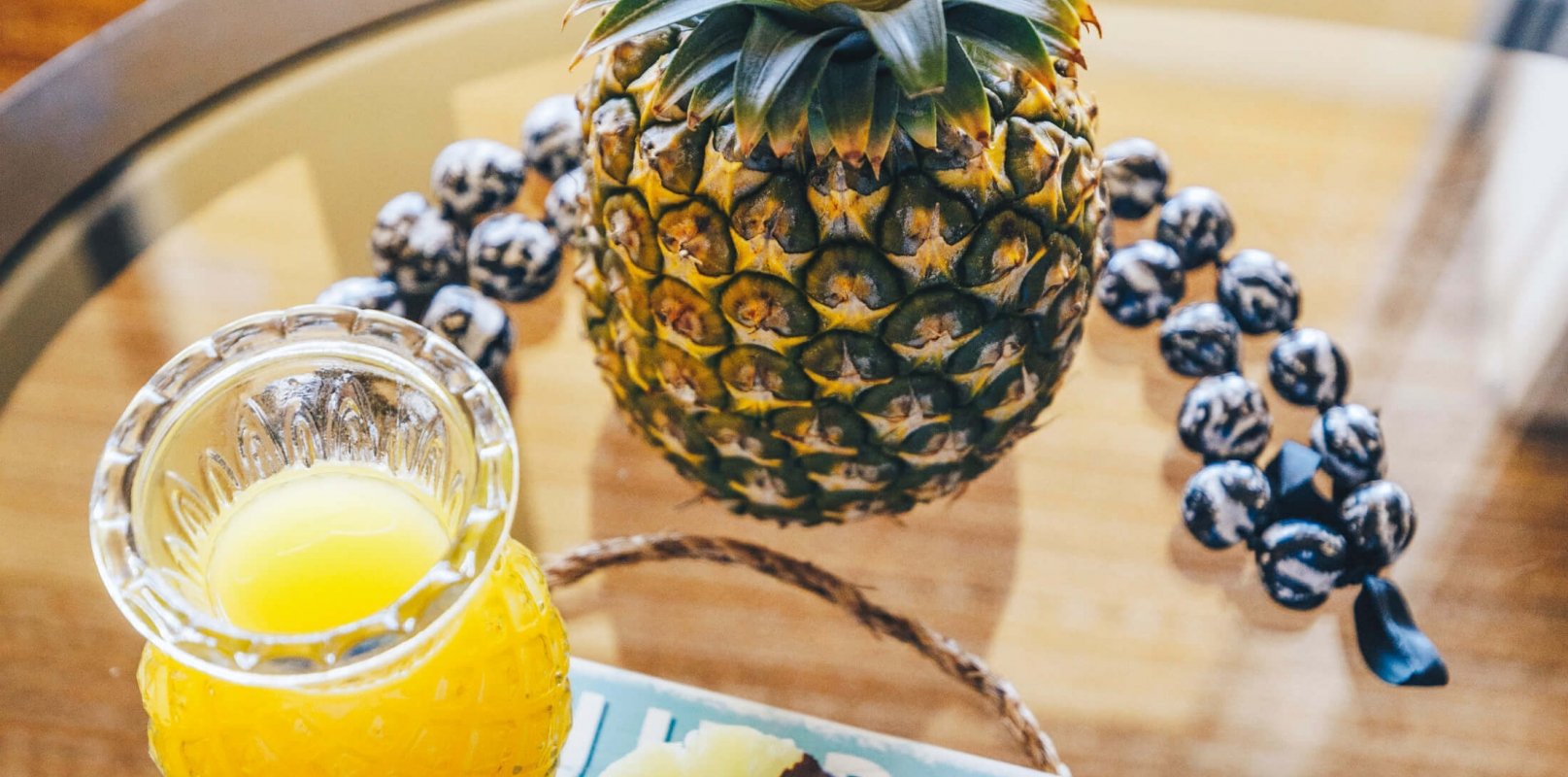 slices of chocolate covered pineapple sit on a blue plate beside a glass of juice and full pineapple on glass table