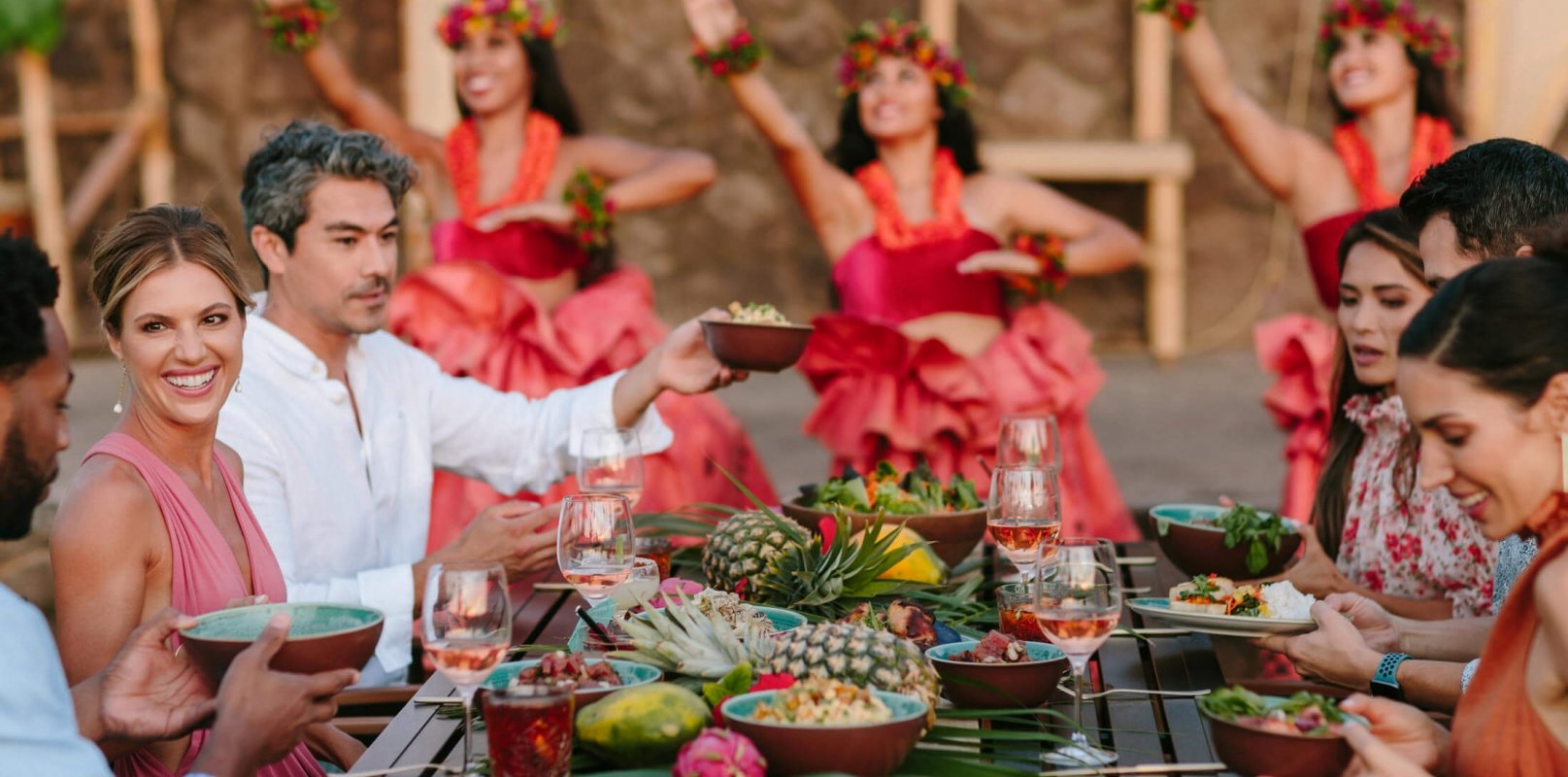 group of people enjoy dinner while women in red perform luau in the background