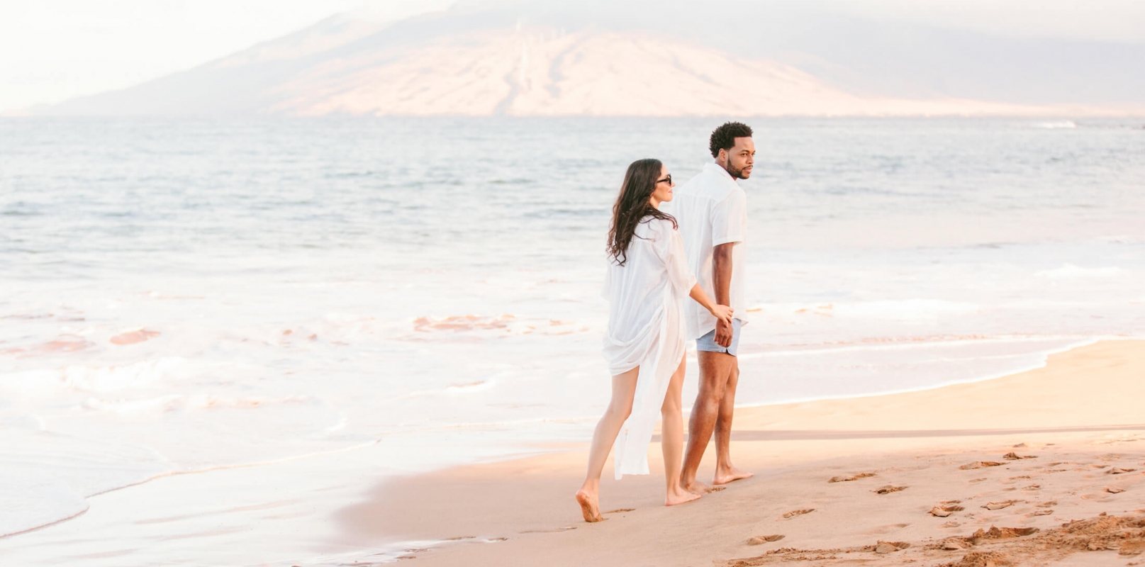 a couple walks along the beach beside the ocean with mountains in the background