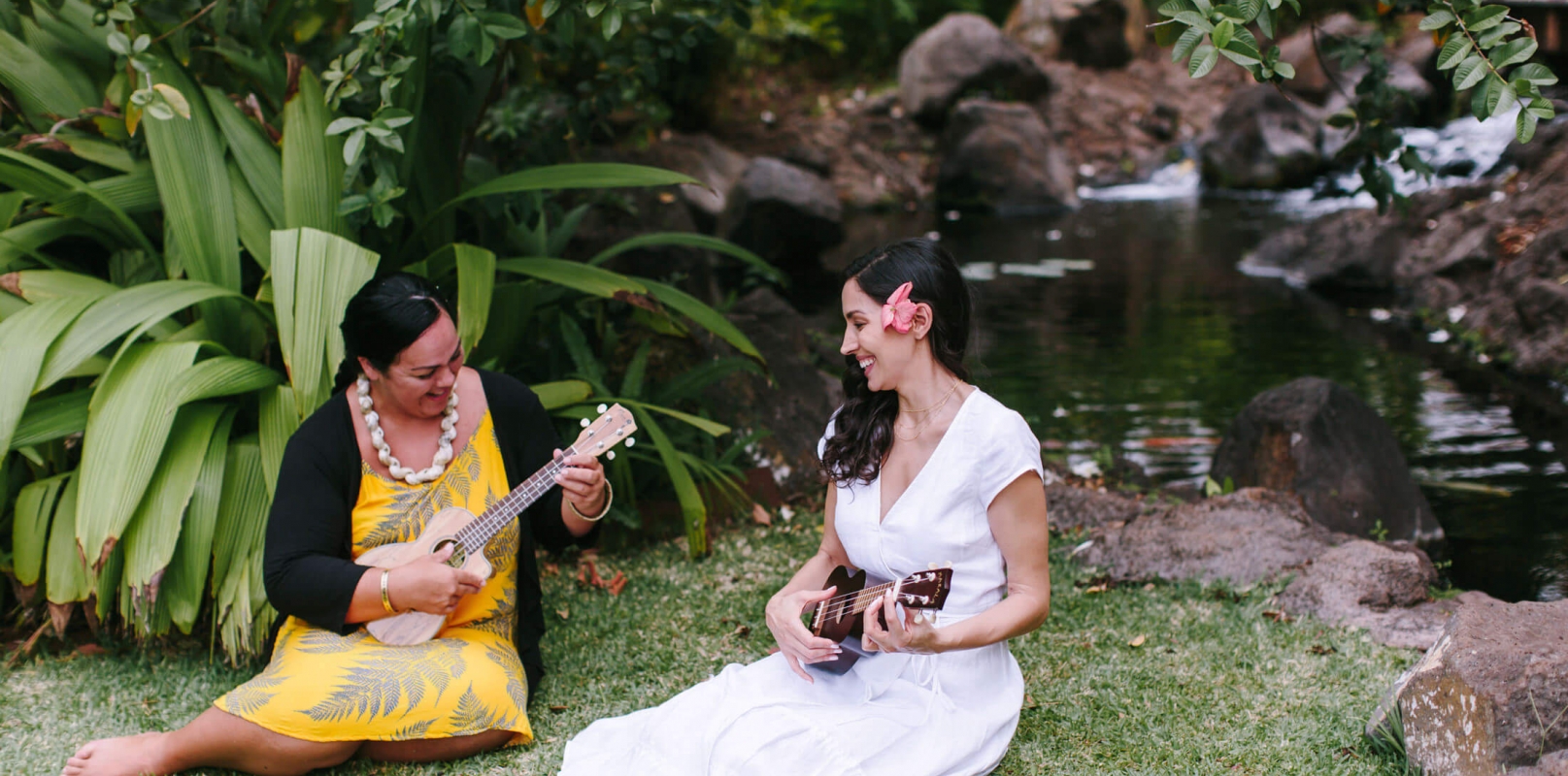 two women sit in the grass while strumming on ukuleles 