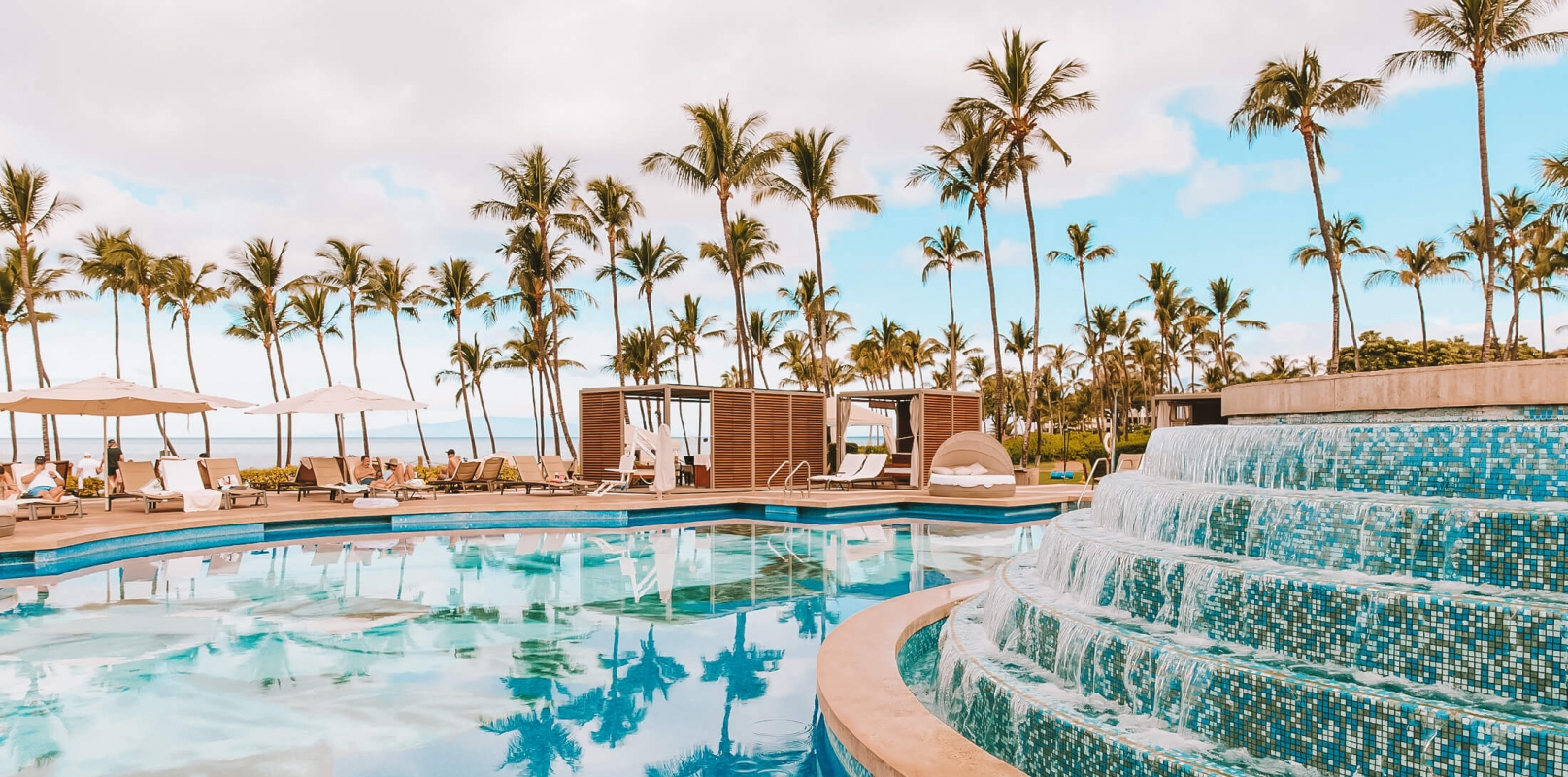 a water feature with multiple levels pours water into a larger pool with palm trees in the background