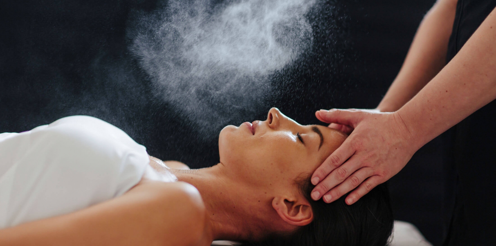 woman lays on her back under a white blanked while her head is massaged and mist is sprayed over her face