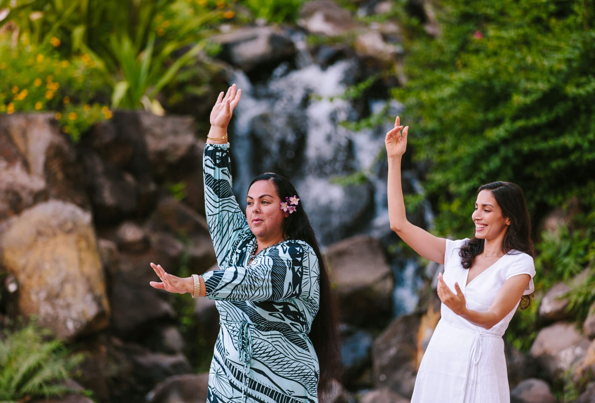 two women hold one hand above their head and one in front of them as they practice dancing in front of.a waterfall feature surrounded by foliage