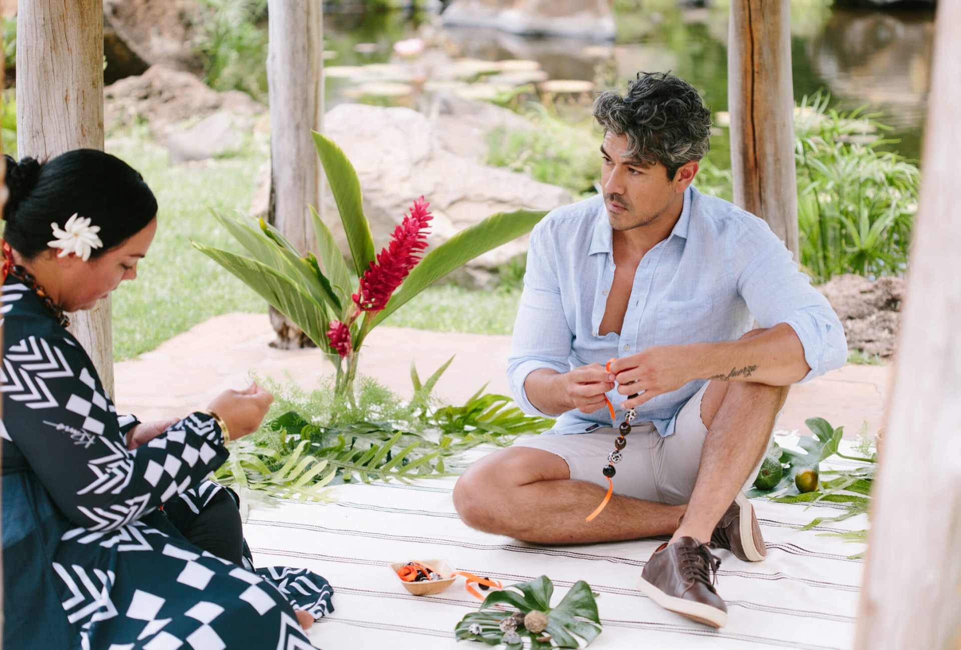a woman in a blue striped dress with a white flower in her hair sits facing a man while weaving a bracelet