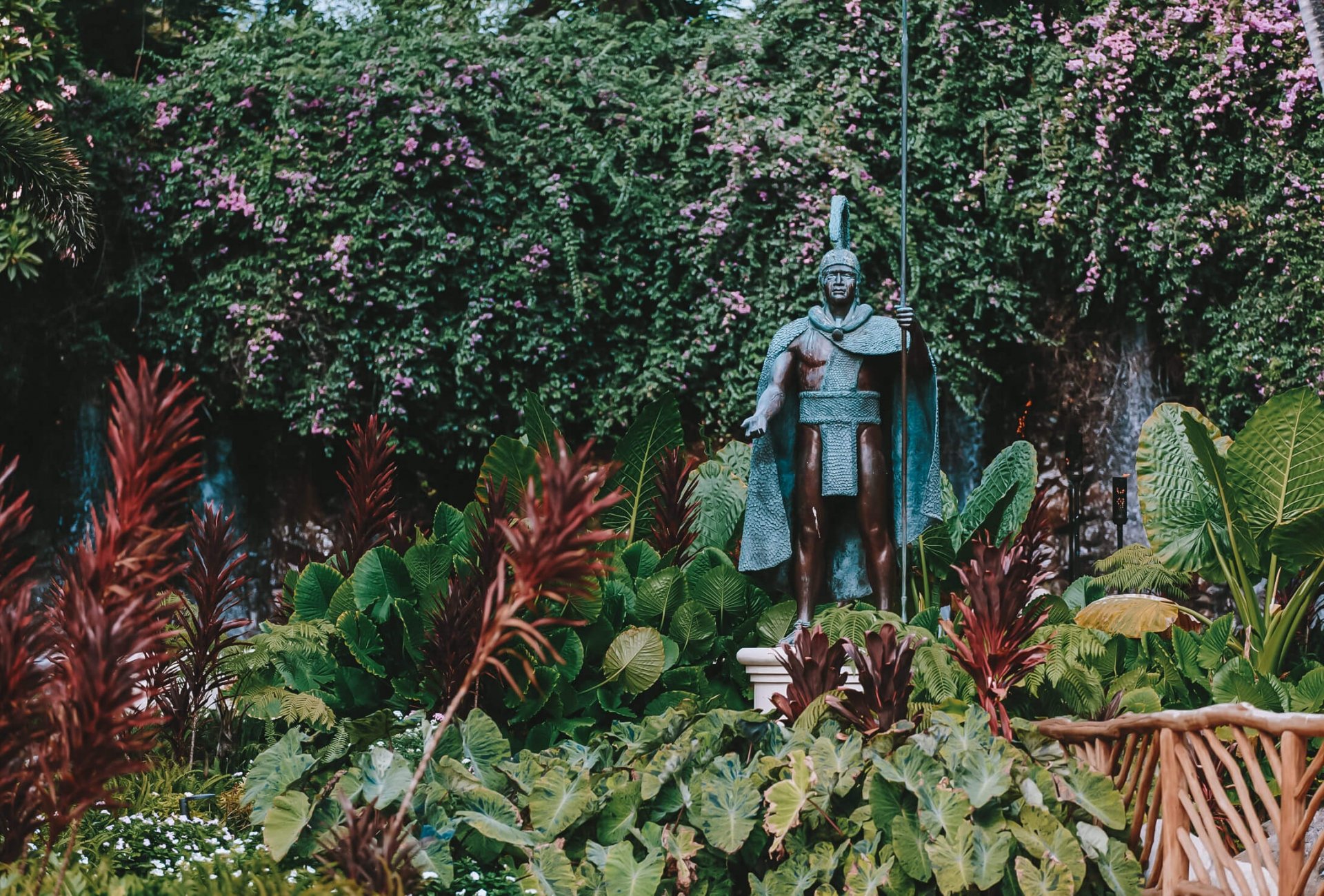 a statue of warrior stands amongst green foliage in front of a forest of trees