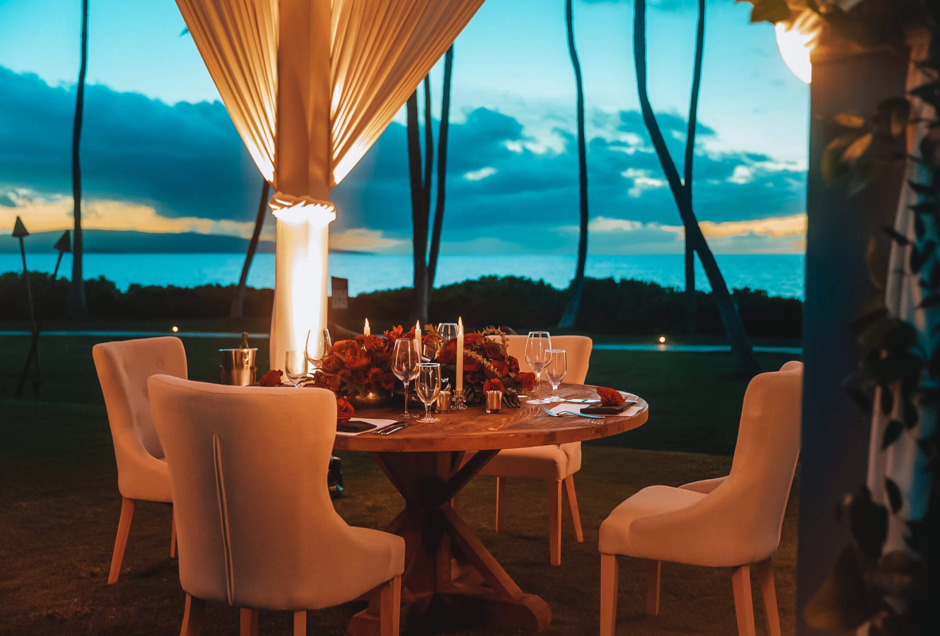 chairs sit around wooden table with white curtains pulled to each corner of pergola is brightly lit against the evening backdrop