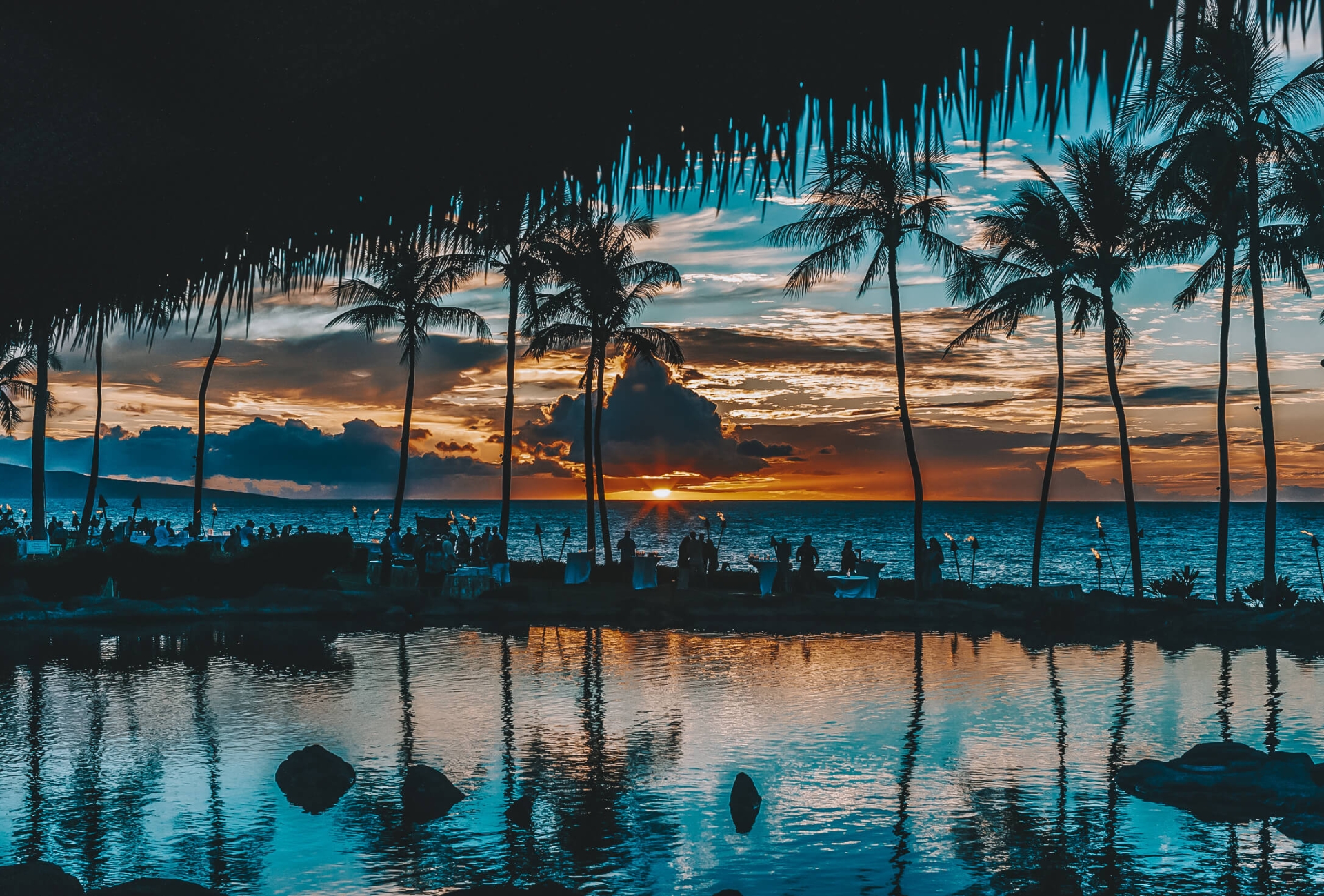 shadow of palm trees reflect off the water in the foreground while the sun sets over the ocean creating an orange and blue sky