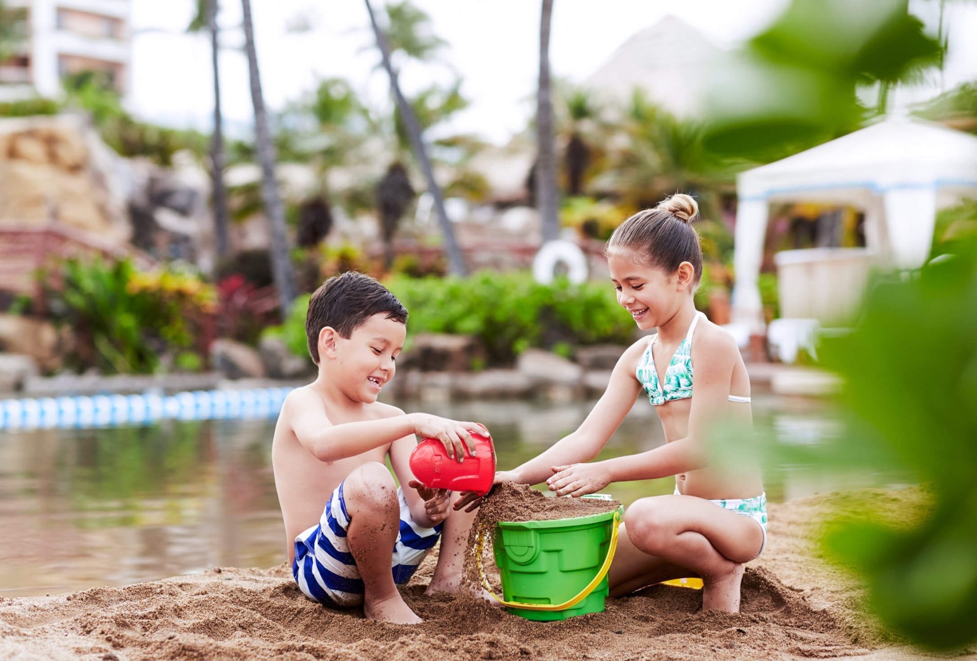 two kids play in the sand with green and red bucket with water in the background