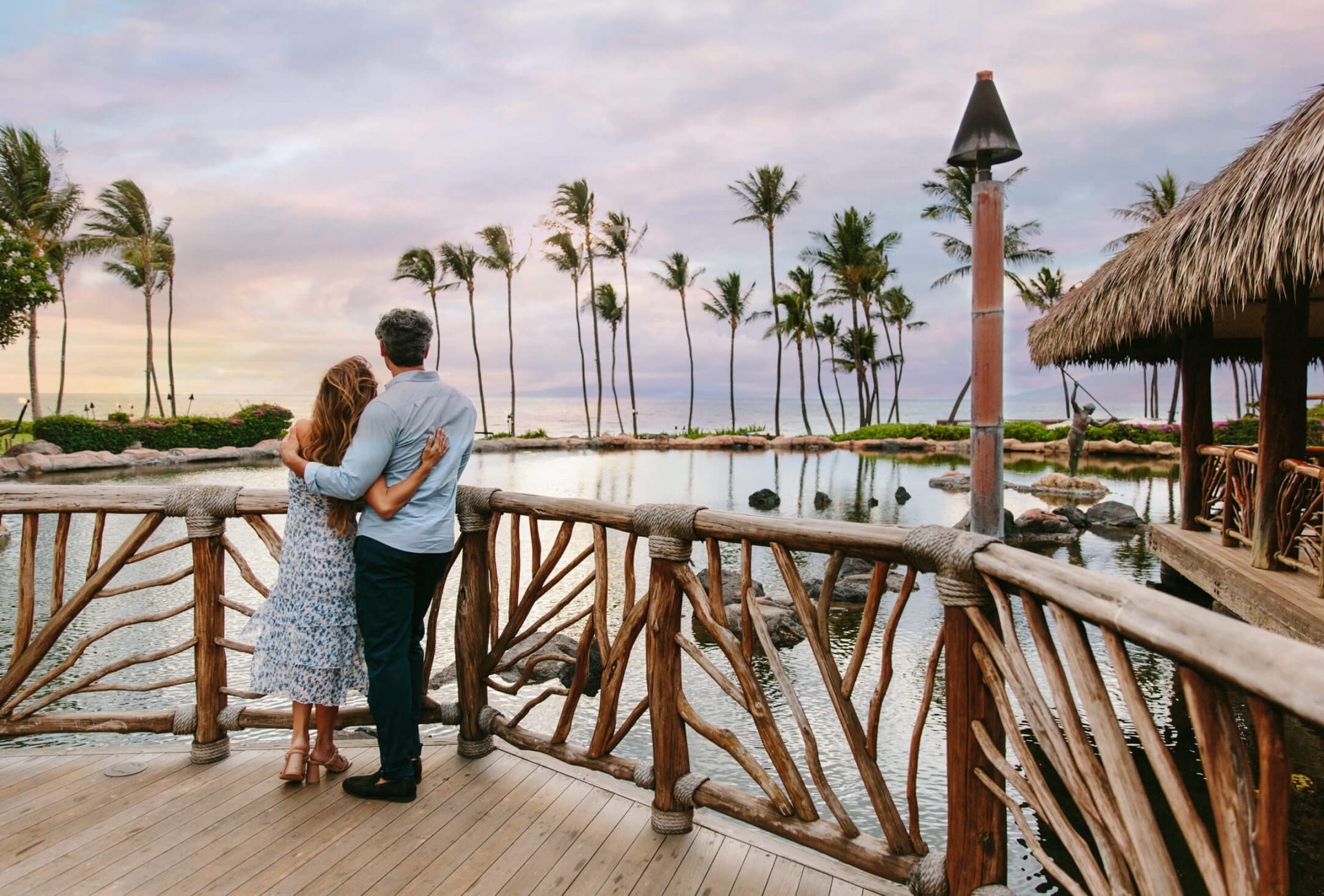 a couple stands on a wooden balcony while looking out at the sun setting