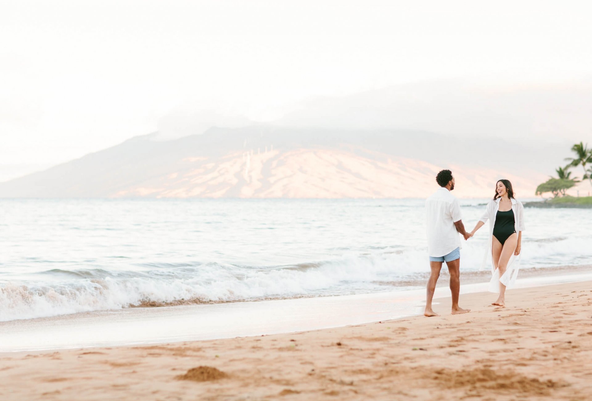 a couple walks along the beach beside the ocean with mountains in the background