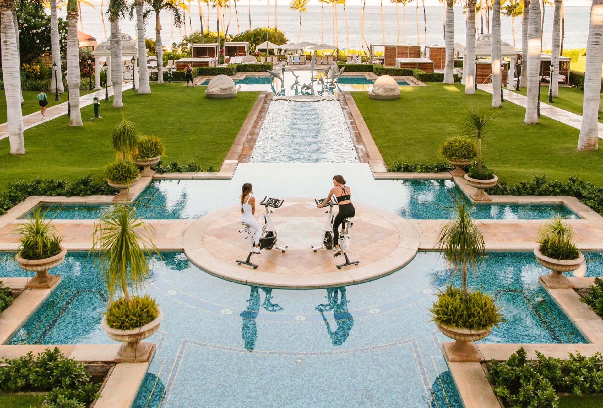 two women wearing black and white workout clothing ride on exercise bikes on platform between pools 