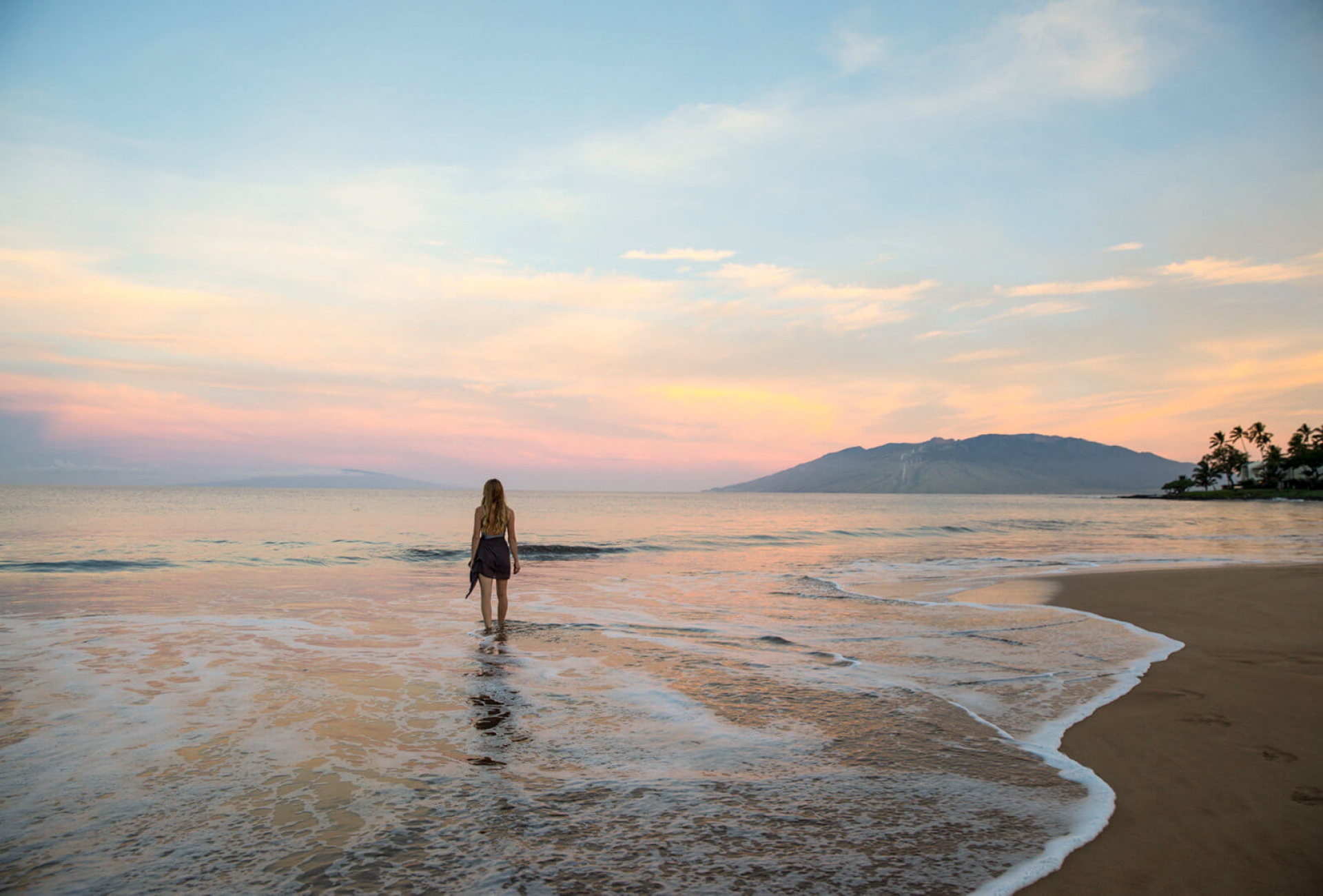 a woman walks toward the ocean on the beach with mountains in the background while the sun sets