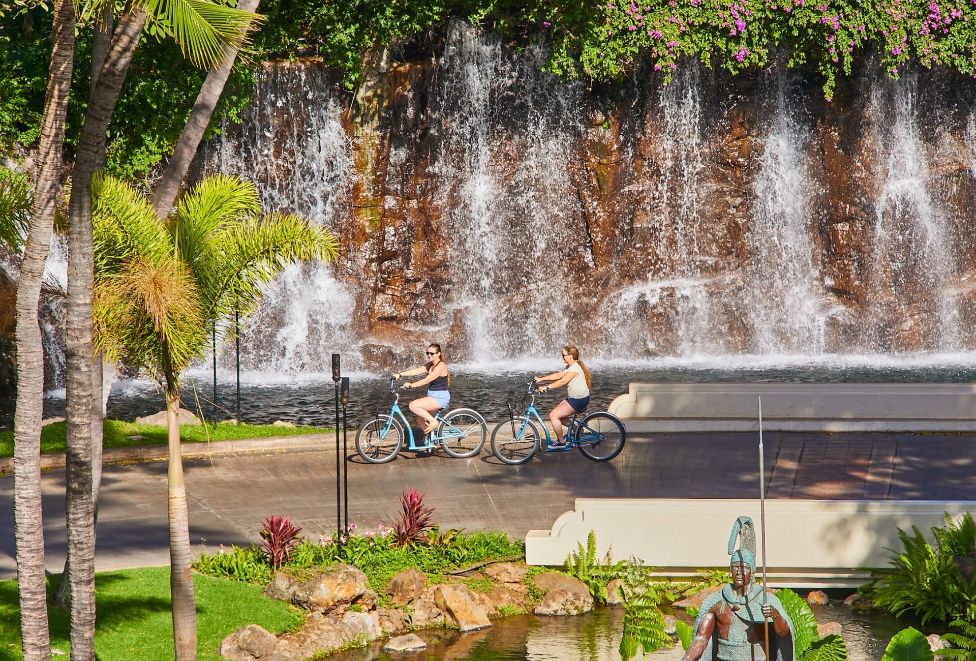 two people ride beach cruiser bikes along a path beside a wall of water
