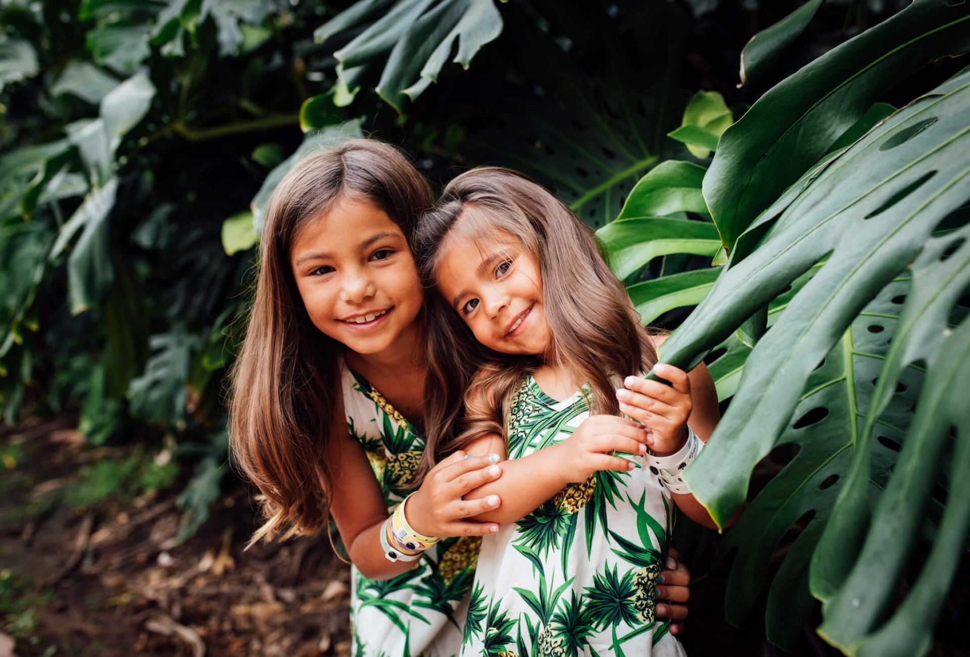 two small children wearing matching green and white outfits stand amongst green leaves of trees 