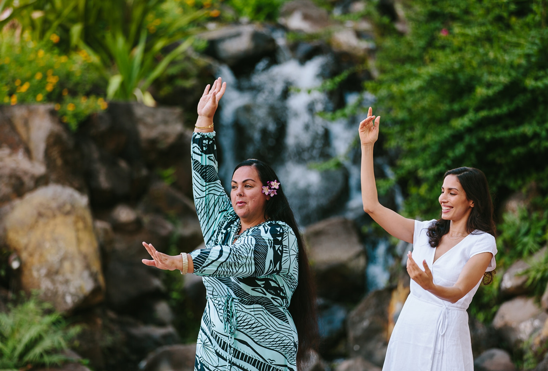 two women hold one hand above their head and one in front of them as they practice dancing in front of.a waterfall feature surrounded by foliage