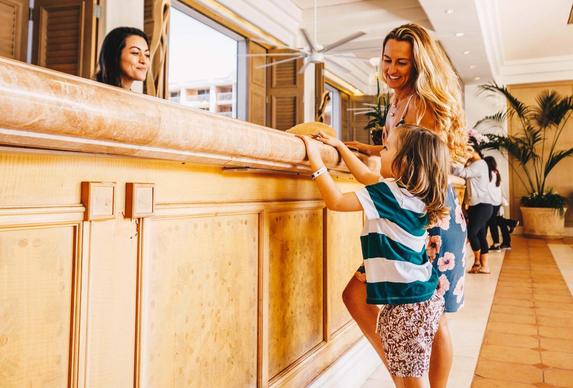 a woman and a child stand on one side of a wooden front desk while the child stands on their toes to see the person on the other side of the desk