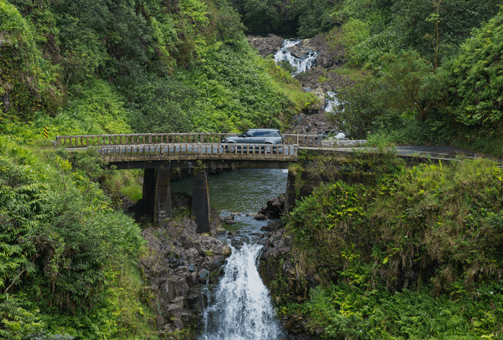 a vehicle drives across a bridge that's situated above a small waterfall and surrounded by green foliage and trees