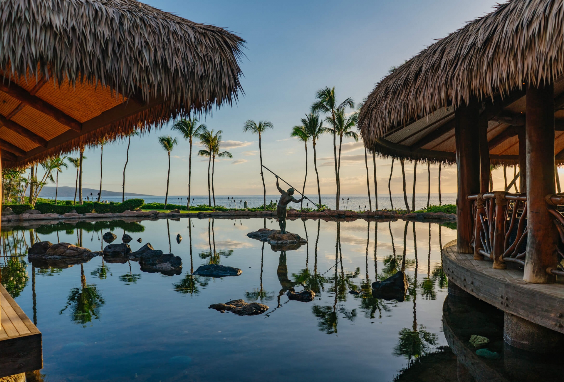 a statue holding a spear stands in the middle of a pond while the sun sets with a hut on either side with blue sky and palm trees in the background