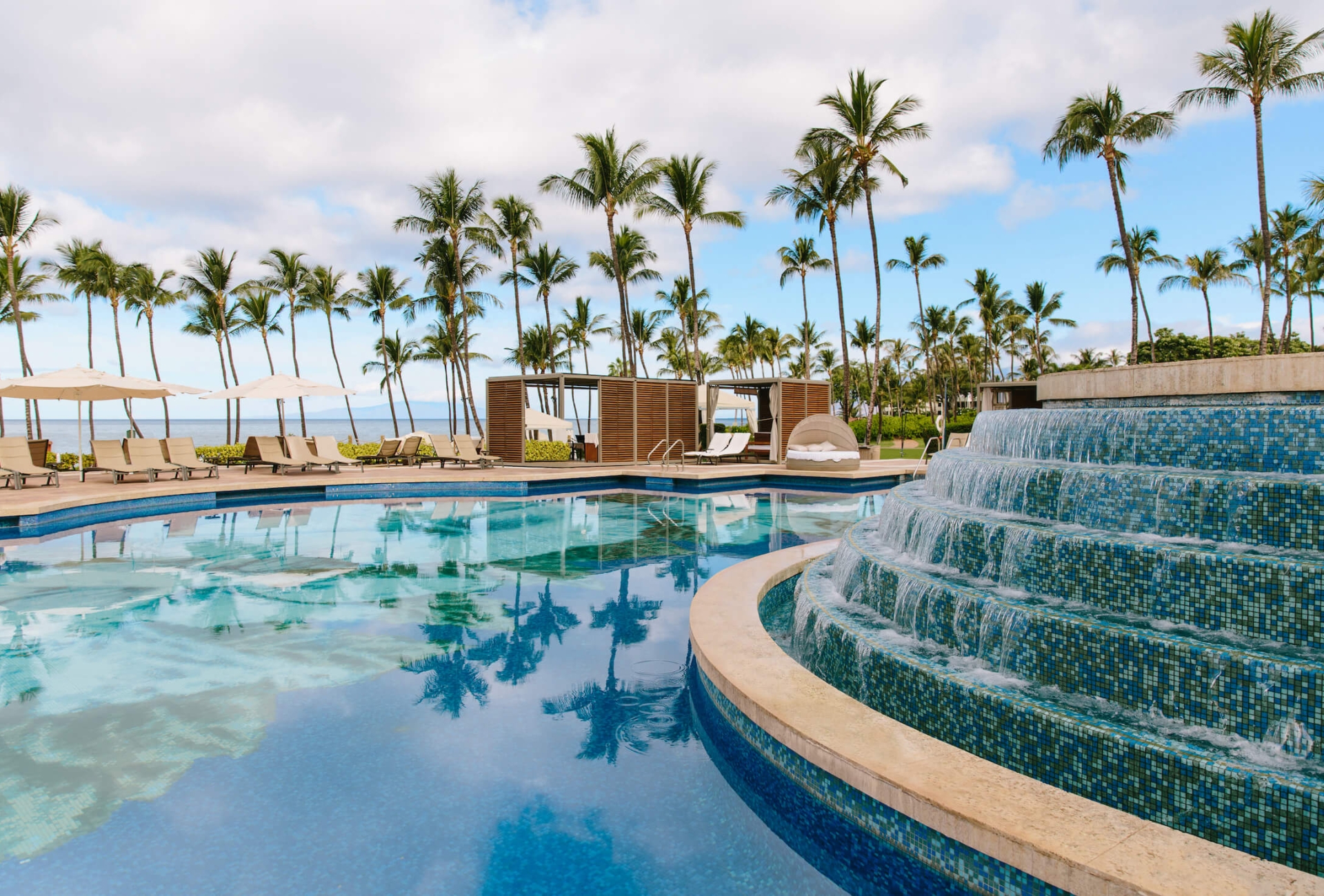 a water feature with multiple levels pours water into a larger pool with palm trees in the background