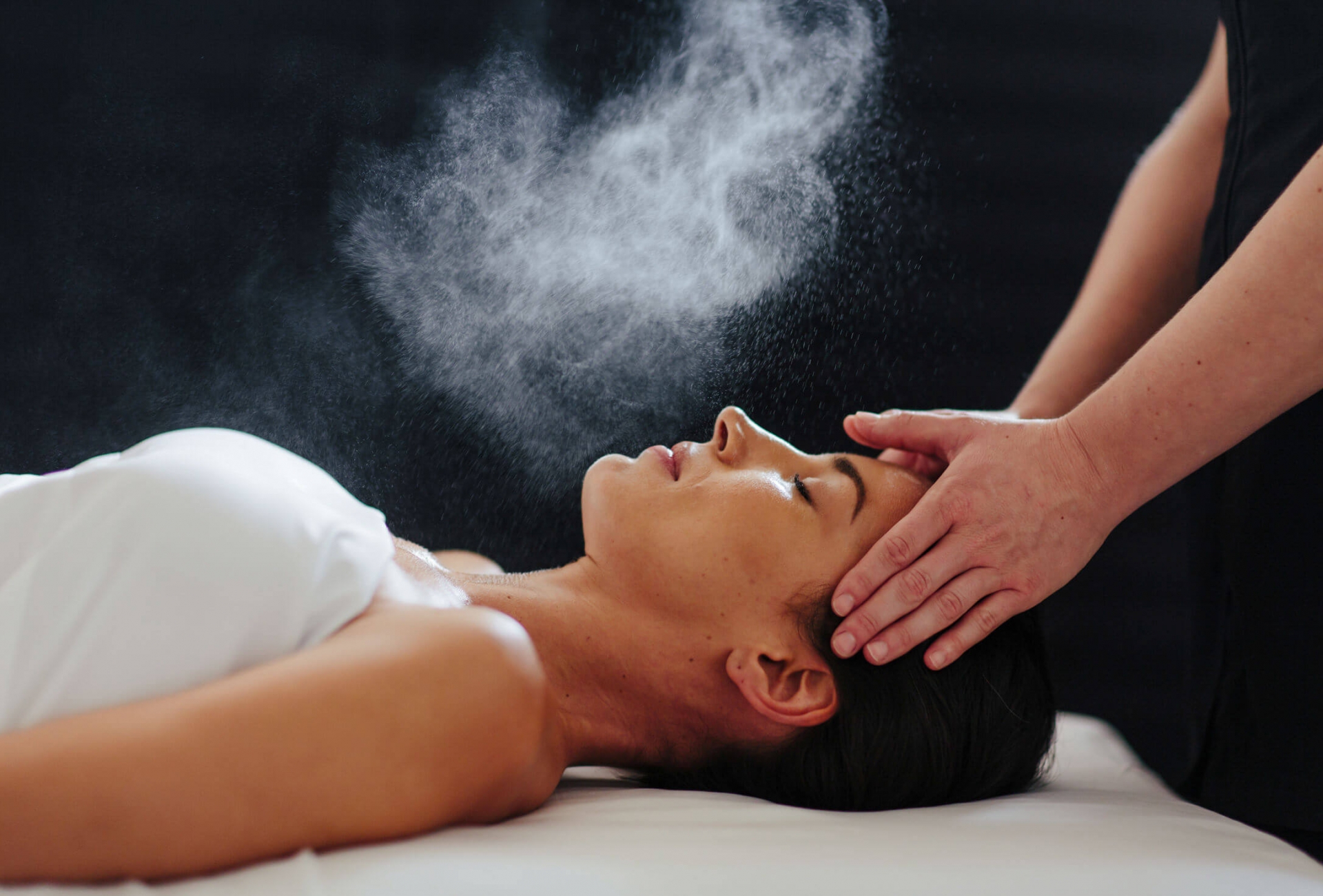 woman lays on her back under a white blanked while her head is massaged and mist is sprayed over her face