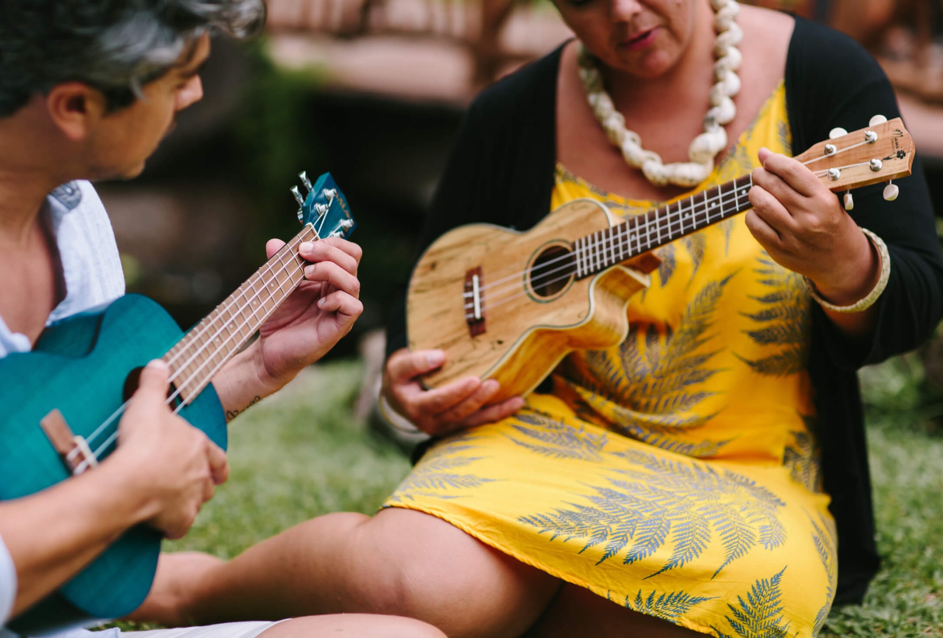 a man and a woman holding brightly colored ukuleles sit on the grass