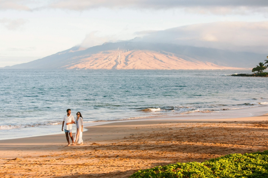 a couple walks along the beach beside the ocean with mountains in the background