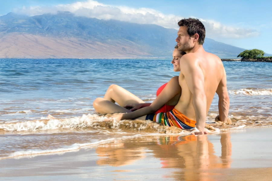 man and child sit on the beach at the ocean's edge with mountains in the background