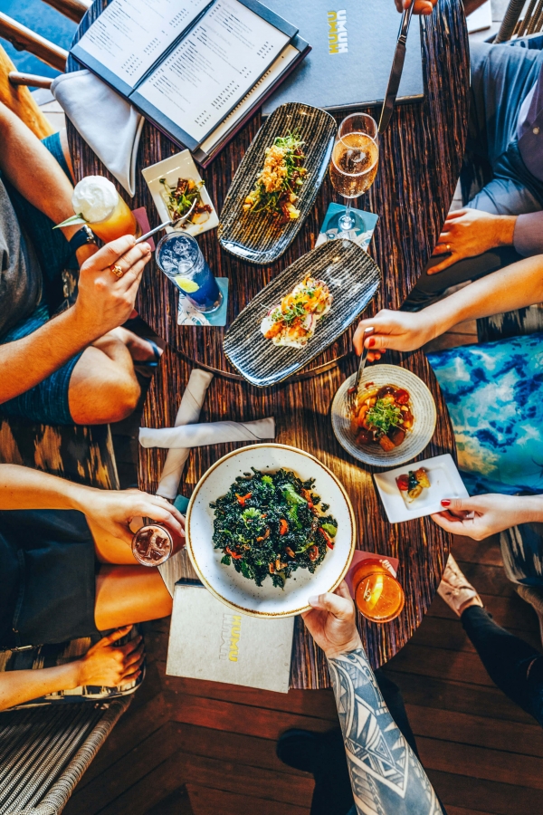 multiple plates of food sit on a wooden table while four people reach over the food with their utensils