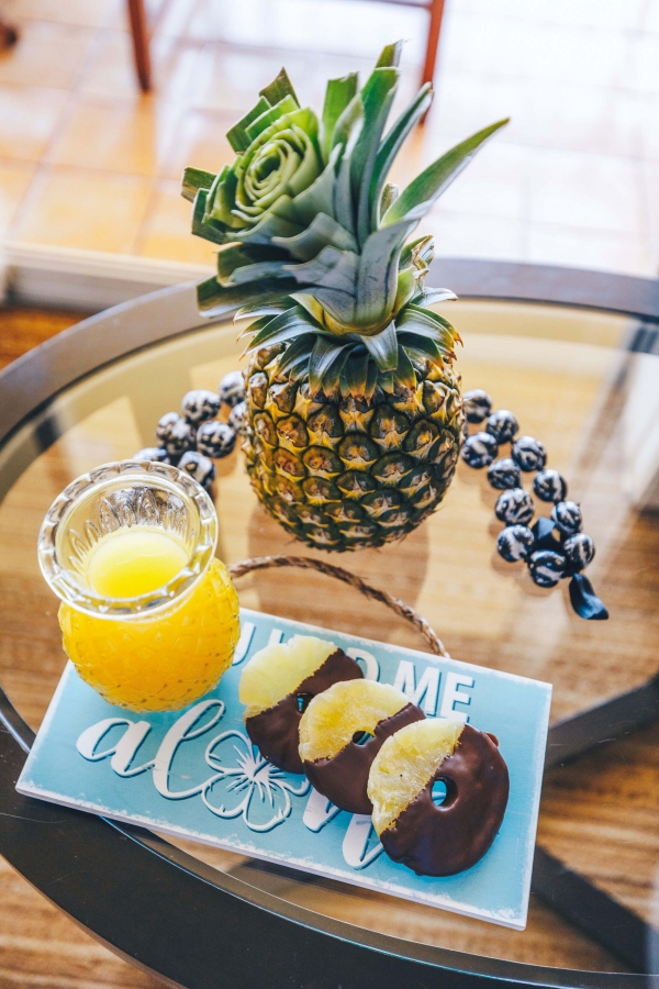 slices of chocolate covered pineapple sit on a blue plate beside a glass of juice and full pineapple on glass table