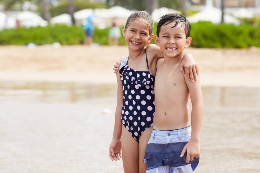 two kids in bathing suits stand on the beach with their arms around each other