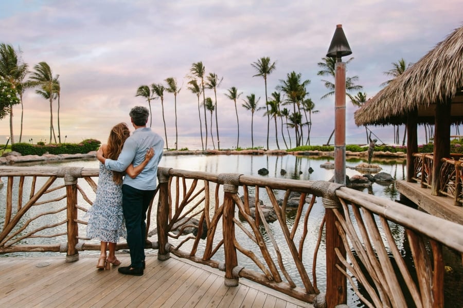 a couple stands on a wooden balcony while looking out at the sun setting