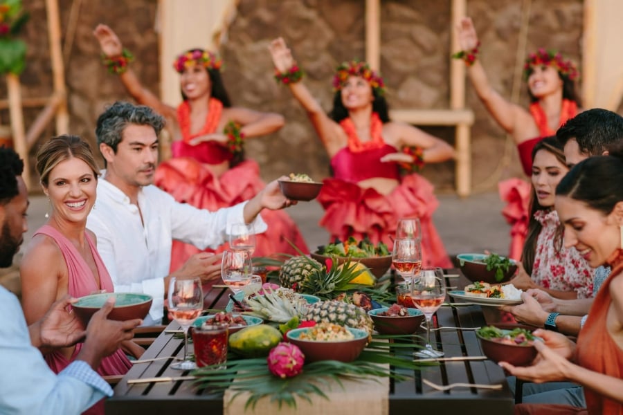 group of people enjoy dinner while women in red perform luau in the background