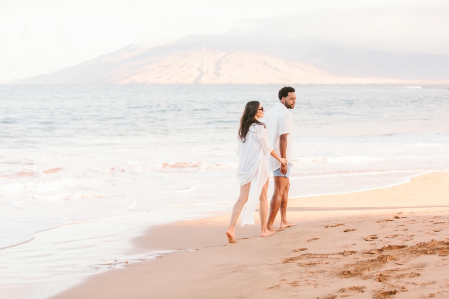 a couple walks along the beach beside the ocean with mountains in the background