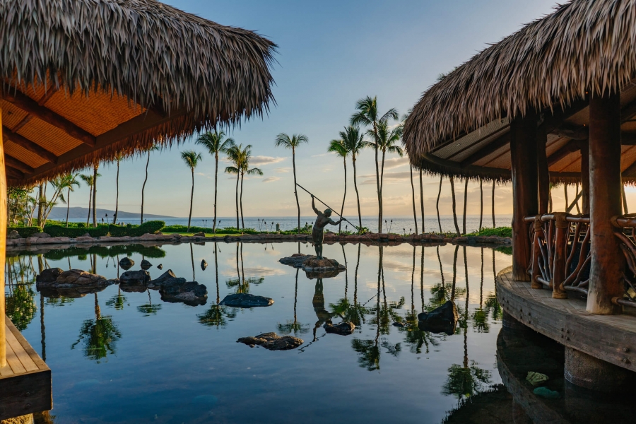 a statue holding a spear stands in the middle of a pond while the sun sets with a hut on either side with blue sky and palm trees in the background