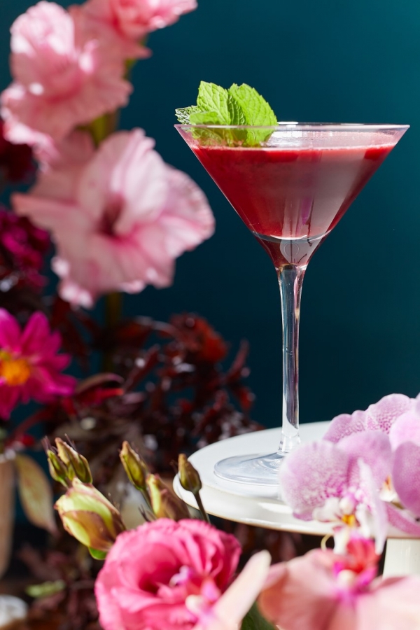 red cocktail in a martini glass sitting beside multiple pink flowers