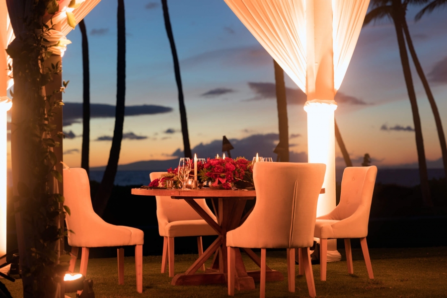chairs sit around wooden table with white curtains pulled to each corner of pergola is brightly lit against the evening backdrop