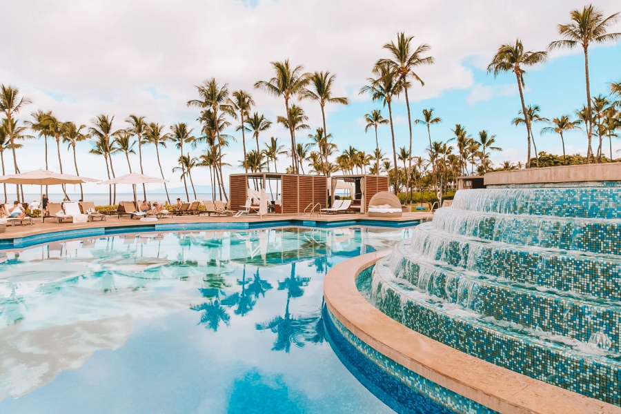 a water feature with multiple levels pours water into a larger pool with palm trees in the background