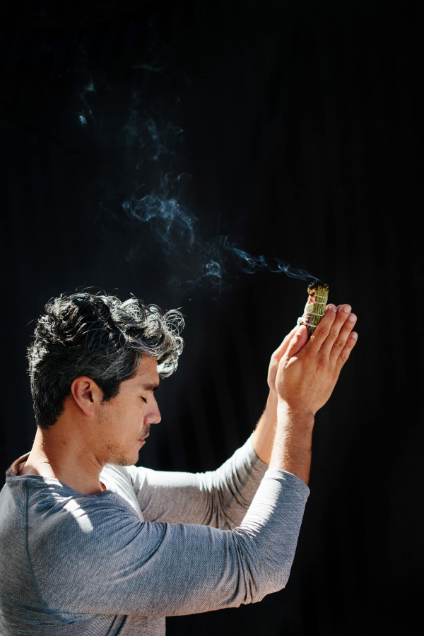 side profile of a man sitting crosslegged on the ground holds his hands up while smoke trails over his head