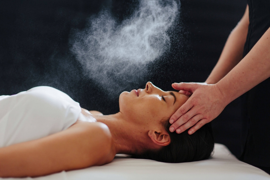 woman lays on her back under a white blanked while her head is massaged and mist is sprayed over her face