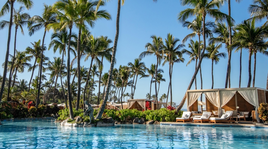 Sun falls on a sparkling blue pool lined with luxury cabanas and waving palm trees